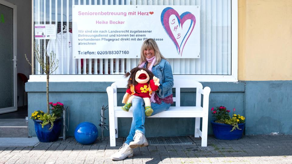 Heike Becker, a middle-aged woman, sitting on a bench with a hand puppet in front of a sign that reads "Seniorenbetreuung mit Herz".