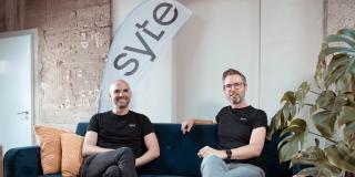 David Nellessen und Matthias Zühlke sitzen auf einer Couch, hinter ihnen eine Fahne ihres Unternehmens syte