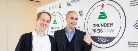 Zwei Männer stehen nebeneinander, der eine zeigt mit dem Daumen auf das Logo vom GRÜNDERPREIS NRW 2020 auf einer Stellwand.