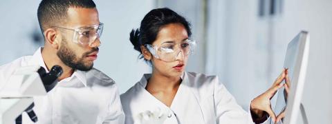 Mann und Frau in Arztkitteln tragen medizinische Schutzbrillen und arbeiten an einem Bildschirm.