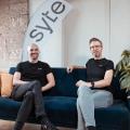 David Nellessen und Matthias Zühlke sitzen auf einer Couch, hinter ihnen eine Fahne ihres Unternehmens syte