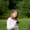Elisabeth Jacobsohn is standing in a park next to a scalpture looking on her smartphone